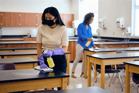 Technicien en entretien ménager nettoyant une salle de classe dans une école, avec des bureaux alignés et un chariot de nettoyage professionnel.